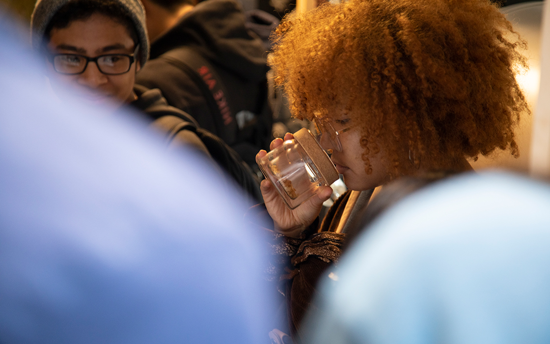UW Tacoma student Elena Mendoza has her nose inside a jar of coffee beans. Mendoza has brightly colored hair and wears glasses. Another student is looking at her. He has on a grey beanie and glasses. In the foreground are the blurred shoulders of other students.