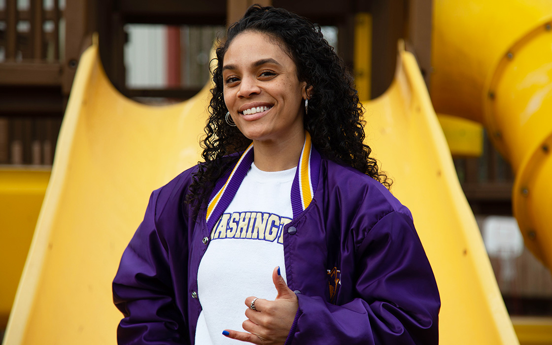 UW Tacoma alumna Amalia Perez stands in front of three yellow slides. She has long, black, curly hair and is wearing a purple and gold Huskies jacket. She also has on a white sweater with the word "Washington" written on it in purple and gold lettering. Perez has her thumb and pinky extended in a "hang loose" symbol.