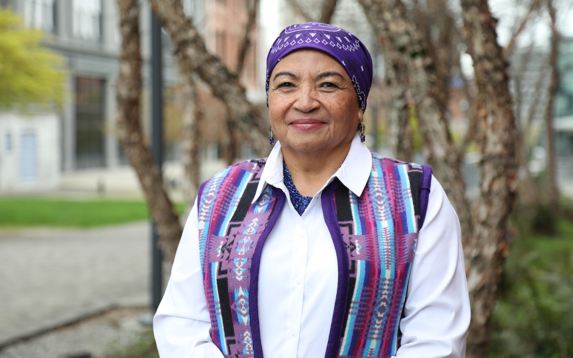 UW Tacoma Ed.D. student Ada McDaniel stands in front of a tree. She is wearing a head wrap that is purple and has symbols woven into it. McDaniel is also wearing  a purple vest with symbols woven into it. She has on a white undershirt.