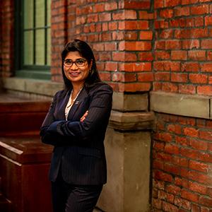 UW Tacoma Professor Divya McMillin poses in front of a red brick wall. She has black hair and is wearing dark colored glasses and a dark colored suit.