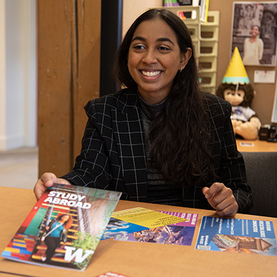 UW Tacoma student Angel Reddy sits at a desk. She is wearing a red stripped jacket and has long black hair. Reddy is smiling. In her right hand she is holding out a copy of a Study Abroad brochure.