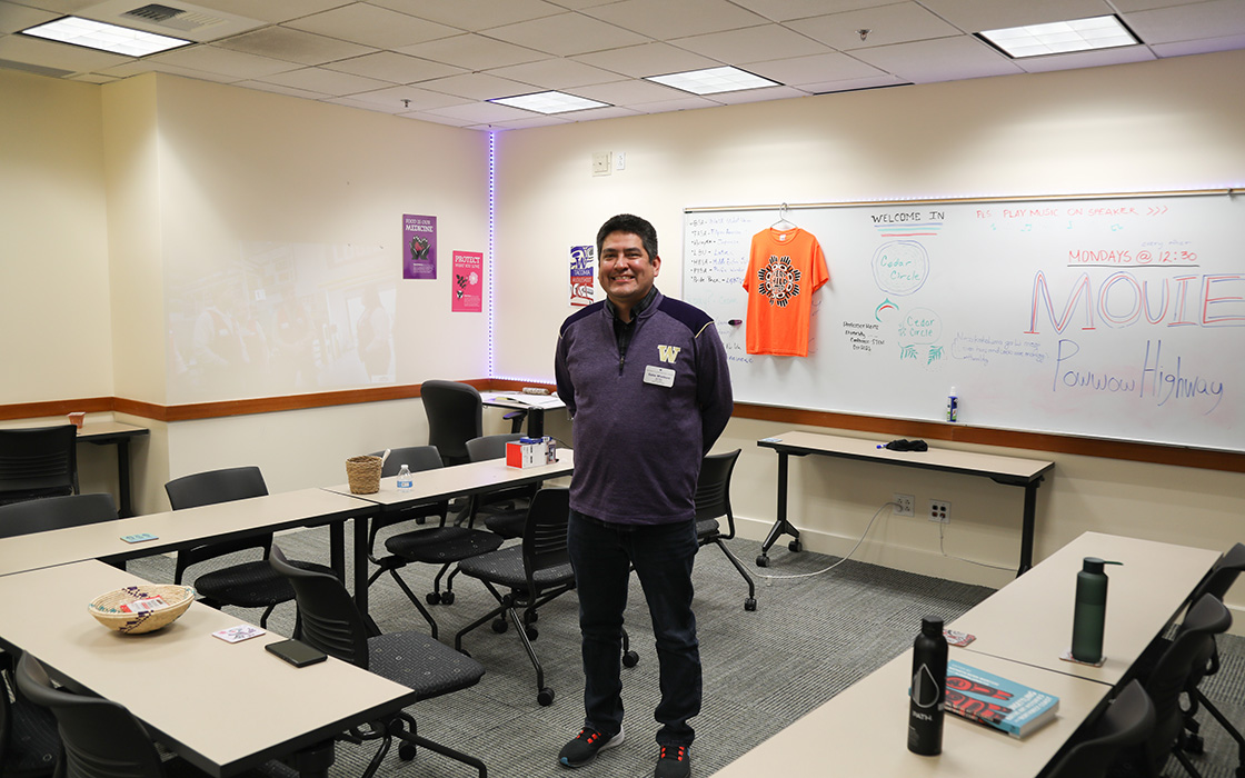 UW Tacoma Tribal Liaison Gabe Minthorn stands in the middle of the Family Room. There are empty tables all around him. There is a whiteboard in the background. There is writing on the board and a shirt hanging off of it by a hanger. 