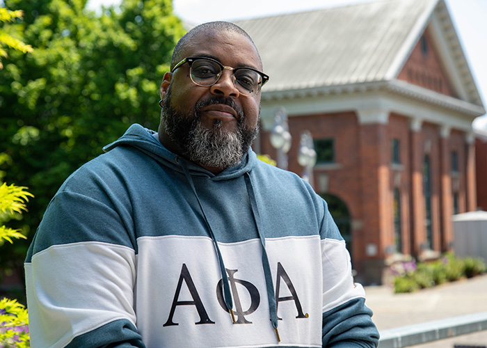 Headshot of UW Tacoma alumnus Jeff Dade. Dade is wearing a green and white sweatshirt with Greek (fraternity) letters on it. He also has on dark colored classes. In the background is a tree with green leaves and the red brick Snoqualmie Power House.