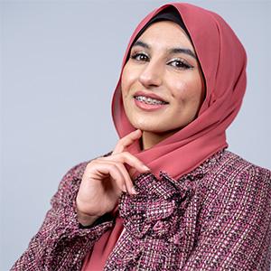 UW Tacoma student Maryam Al Darraji poses against a blank backdrop. She is wearing a pink headscarf and a plaid suit. Her right hand is held up close to her face with her right index finger touching her chin.