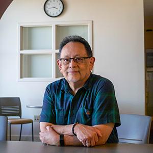 UW Tacoma faculty member Ronnie San Nicolas sits at a desk, his arms crossed in front of him. He is wearing a dark colored, striped shirt. San Nicolas is wearing glasses and has a goatee. In the background there is a white wall, a window and a clock.