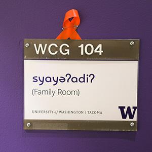 A white sign against a purple wall. The sign reads WCG 104. It says syayəʔadiʔ  underneath which translates in Lushootseed to Family Room.