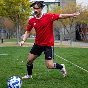 Student Martin Shehata is about to kick a soccer ball. He is wearing a red shirt and black shorts. He has on white cleats and dark socks. His dark colored hair is moving up in the photo. In the background are buildings, trees and grass.