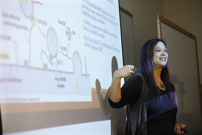 Image: Assistant Professor, Dr. Anna Groat-Carmona, stands smiling in front of a screen while leading a biology class.