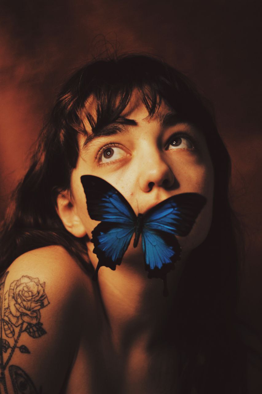 A portrait of a woman, viewed at an angle and cropped to her head and upper shoulders, looks to the upper left of the frame. A blue butterfly sits on her cheek, obscuring her mouth and chin.