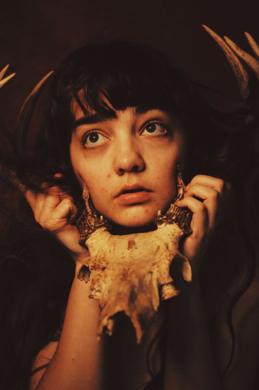 A portrait of a woman holding the skull of an animal by its antlers so that it rests underneath her chin. She is looking to the upper left of the frame.