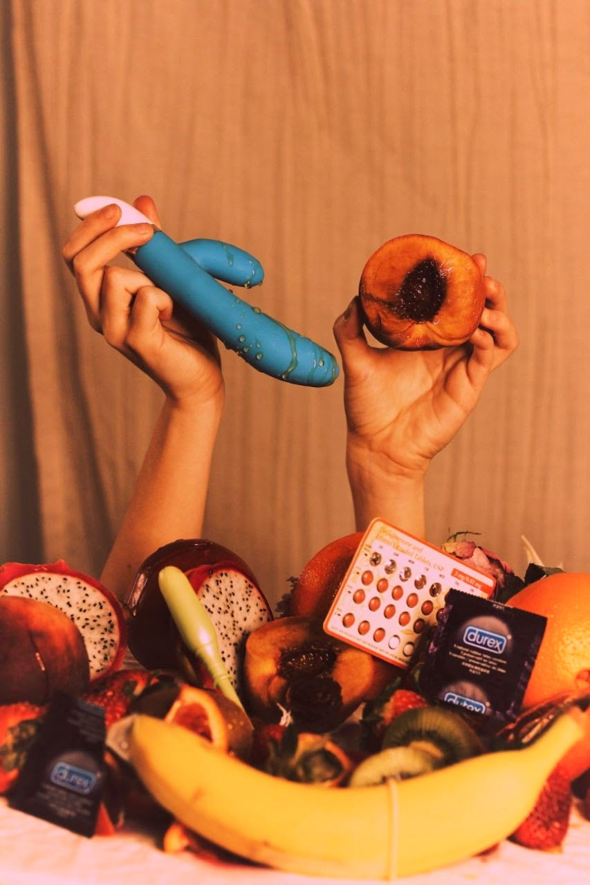 IMAGE: A table is covered in bananas, plums, oranges, and other indistinct produce, as well as birth control pills and condoms. A pair of hands reach up from behind the table, holding a halved plum and a sex toy covered in the plum's juices.