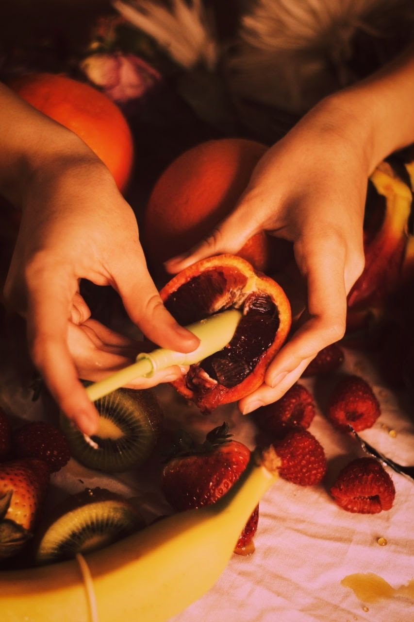 IMAGEL: Two hands extend from out of frame. One hand holds a halved fruit while the other inserts a tampon applicator into the fruit's open face. Underneath the hands, a table is strewn with bananas, kiwis and strawberries.