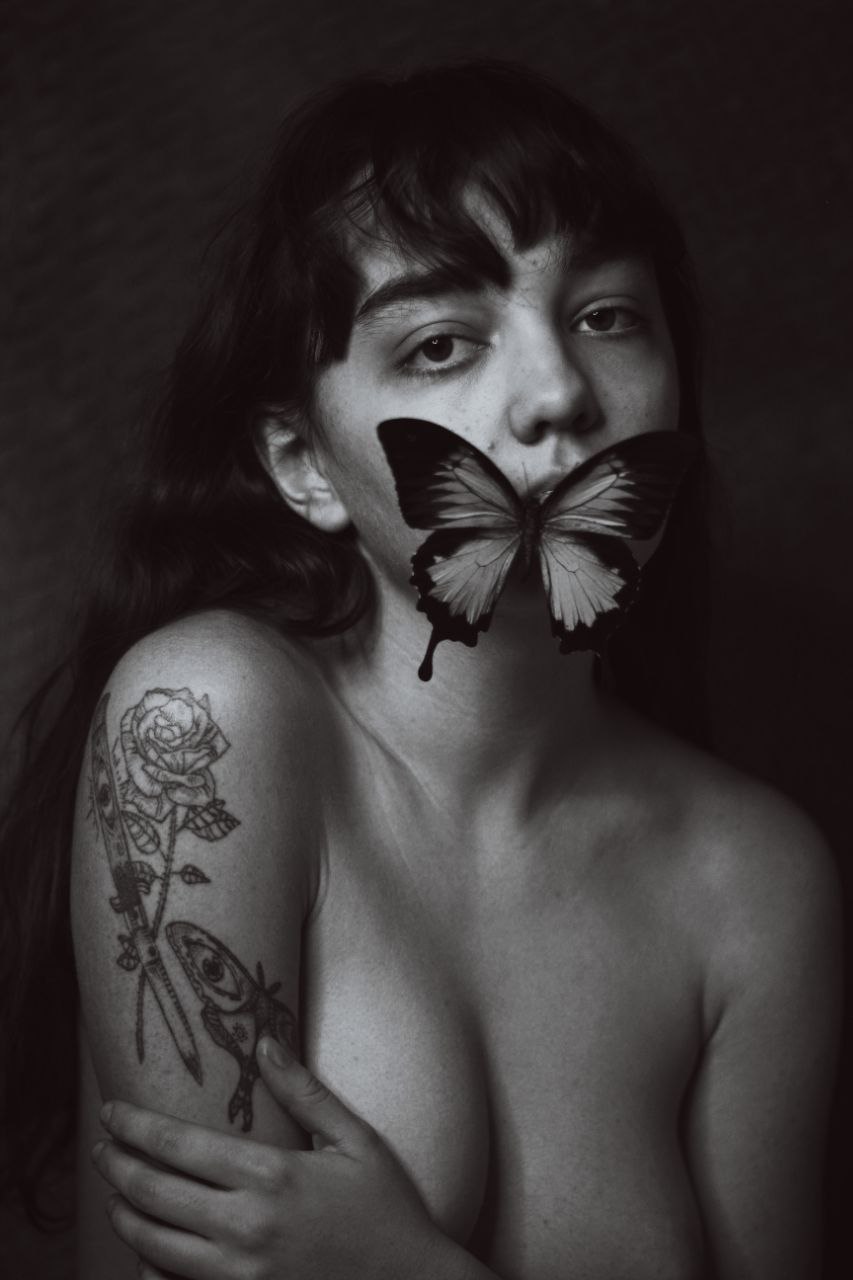 A portrait of a woman from the upper mid-torso up. She has her arms crossed and is looking directly at the camera. A butterfly resting on her cheek obscures her mouth and chin.