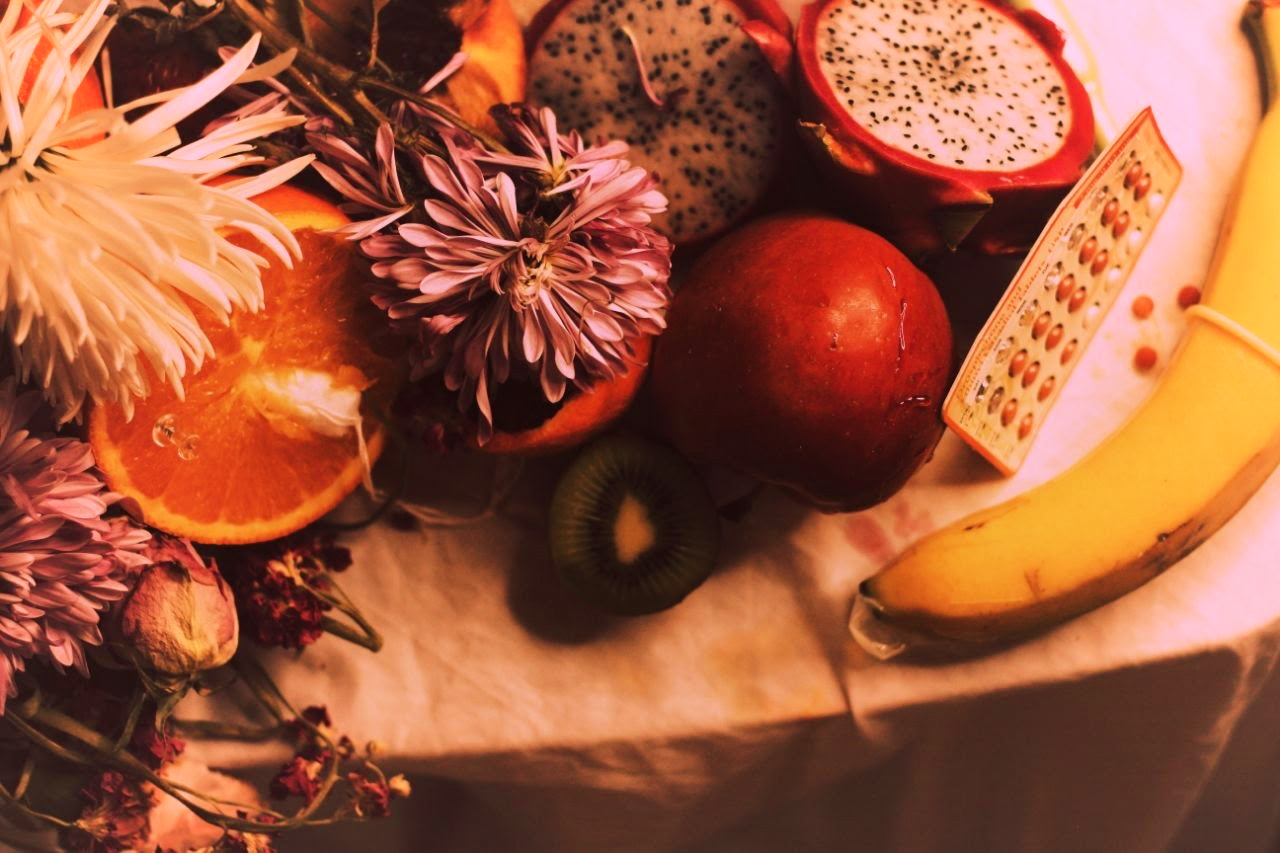 Image: A table is decorated with flowers, a halved orange with an inserted tampon, a halved kiwi, a whole plum, a packet of birth control pills as well as three loose pills on the table surface, and a banana sheathed in a condom.