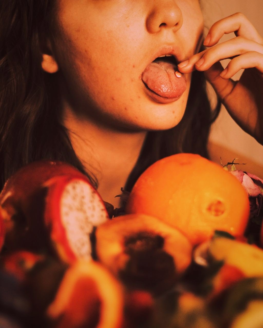 Image: A woman visible from nose to neck places a pill on her tongue. In the foreground, various fruit are strewn on a flat surface.