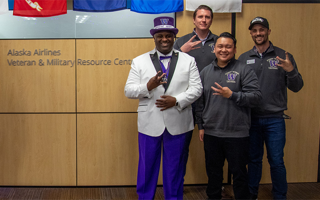 UW Tacoma alumnus Mark Glenn in purple top hat, white jacket, purple pants, posing with staff members from the Alaska Airlines Veteran & Military Resource Center