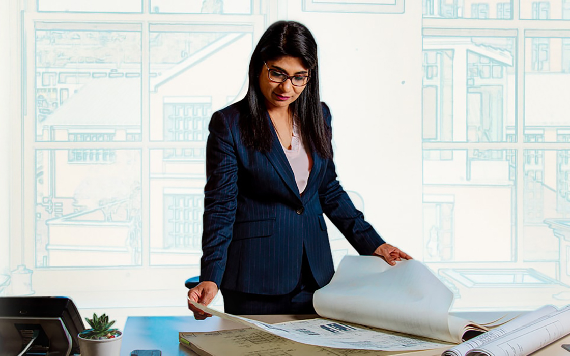 UW Tacoma professor looks over a series of blueprints on her desk. There is an artistic rendering of blueprints in the background.