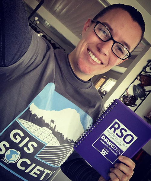 A selfie of Ashley Young. She is wearing a GIS Society t-shirt and is holding a blue copy of the university RSO guidebook. She is wearing glasses and her brown hair is pulled back in a ponytail.