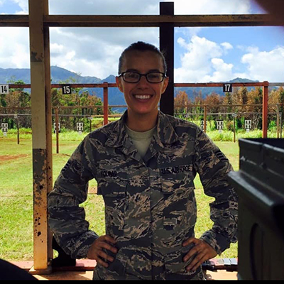 UW Tacoma student Ashley Young poses for a picture in her military uniform. The uniform is camouflaged brown and black. Ashley has her brown hair pulled back in a ponytail and is wearing glasses. 