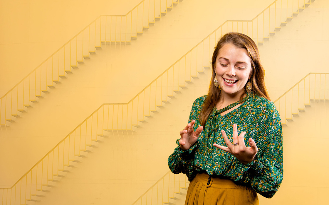 UW Tacoma alumna Abigail Lawson stands in front of a gold colored background. The background has three staircases on it. Lawson has long brown hair and is wearing a dark, floral print shirt and orange pants.