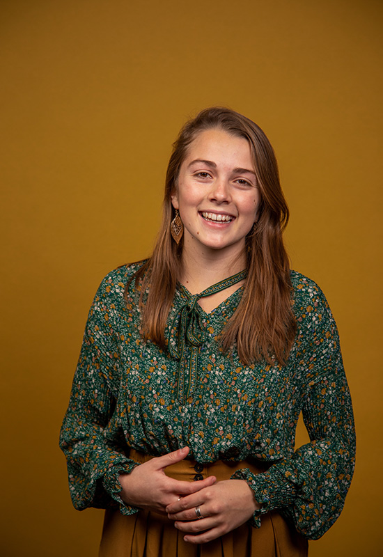 UW Tacoma alumna Abigail Lawson stands in front of a gold colored background. She is wearing a dark colored, floral print blouse and orange pants.
