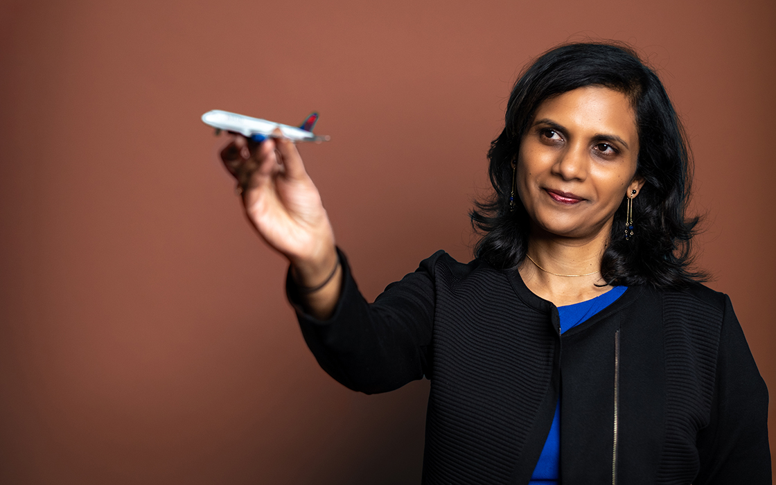 UW Tacoma faculty member Menaka Abraham stands in front of a brown backdrop. Abraham is wearing a black coat with a blue blouse underneath. She is holding a small toy airplane in her right hand which is extended toward the camera.