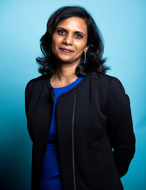UW Tacoma faculty member Menaka Abraham stands in front of a blue background. Abraham is wearing a black coat with a blue blouse underneath. Her arms are behind her back.