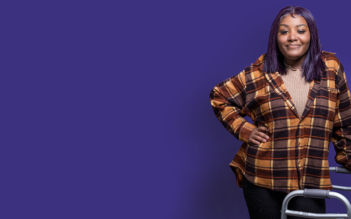 UW Tacoma student Chanise Jackson stands against a purple background. She is wearing a plaid shirt and his holding onto a walker.