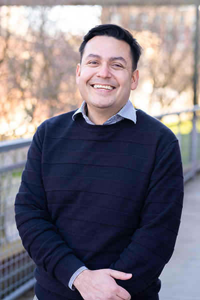 UW Tacoma faculty member Rubén Casas. Casas is wearing a black sweater and has black hair. There are leafless trees in the background.