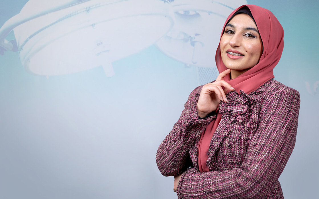UW Tacoma student Maryam Al Darraji poses in front of a background of surgical lights. She is wearing a plaid suit and a red headscarf. Her left hand is folded across her stomach and her right hand is extended toward her chin with one finger touching her chin.