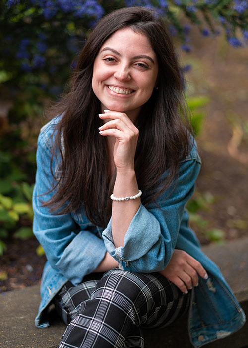 UW Tacoma senior Leticia Barreto sits on a concrete bench. Her left arm is propped up with her hand under her chin. She is wearing striped pants and a blue denim shirt. She has long black hair.