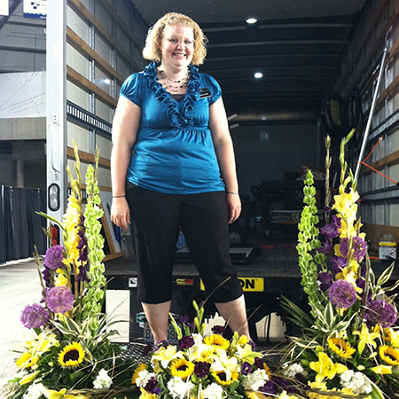 UW Tacoma staff member BrieAnna Bales. Bales is standing on the lift gate of a delivery truck. There are bouquets of flowers at her feet of varying colors. Bales is wearing black capri pants and purple blouse. She was short dirty blond hair and is wearing glasses.