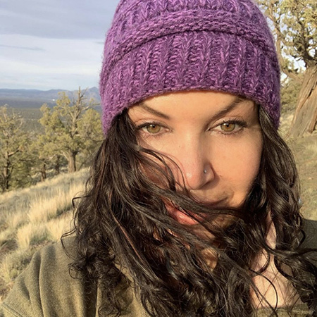 Headshot of UW Tacoma staff member Isabella Webb. Webb is wearing a light blue knitted cap. Her long, brown hair covers part of her face. In the background are rolling hills and tres.