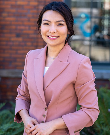 Headshot of UW Tacoma faculty member Sunny Cheng. She  has long black hair pulled back in a ponytail. She is wearing a pink coat. Behind her are some bricks from the Snoqualmie Library.