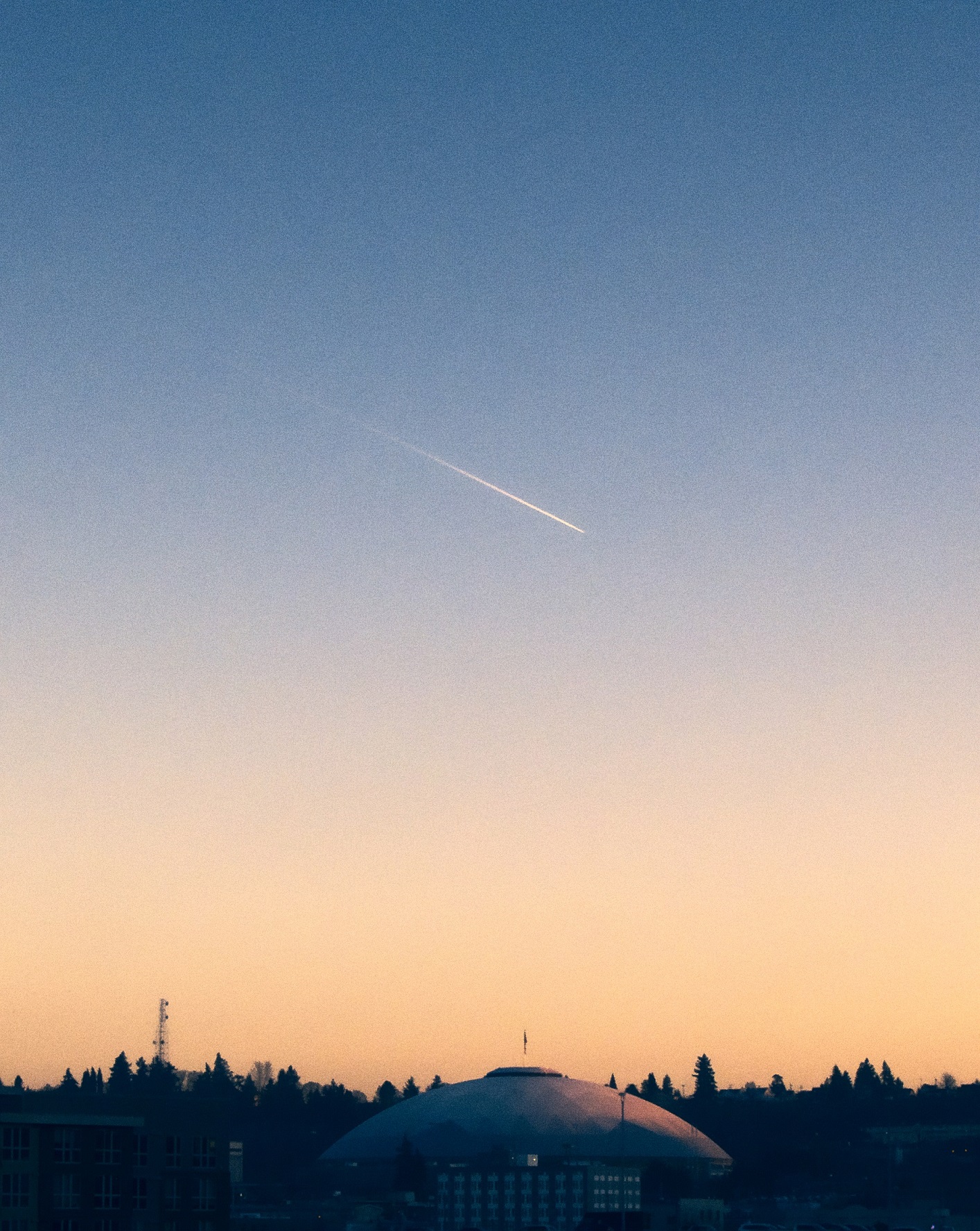 PICTURED: A horizontal photograph of the Tacoma Dome and the sky at sunset, with the latter taking up the upper four fifths of the image. The vapor trail of an air craft is in the sky.