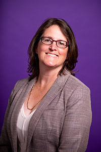 An image of Darcy Janzen, a white woman with shoulder-length brown hair, wearing a blazer and glasses, smiling at the camera, on a purple background.