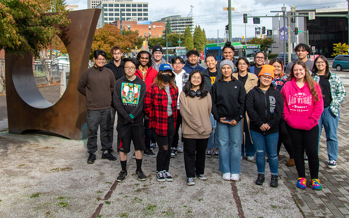 Group of UW Tacoma Asian American and Pacific Islander students standing alongside bronze sculpture Maru, by Gerard Tsutakawa, with railroad tracks of Prairie Line Trail emerging in foreground.
