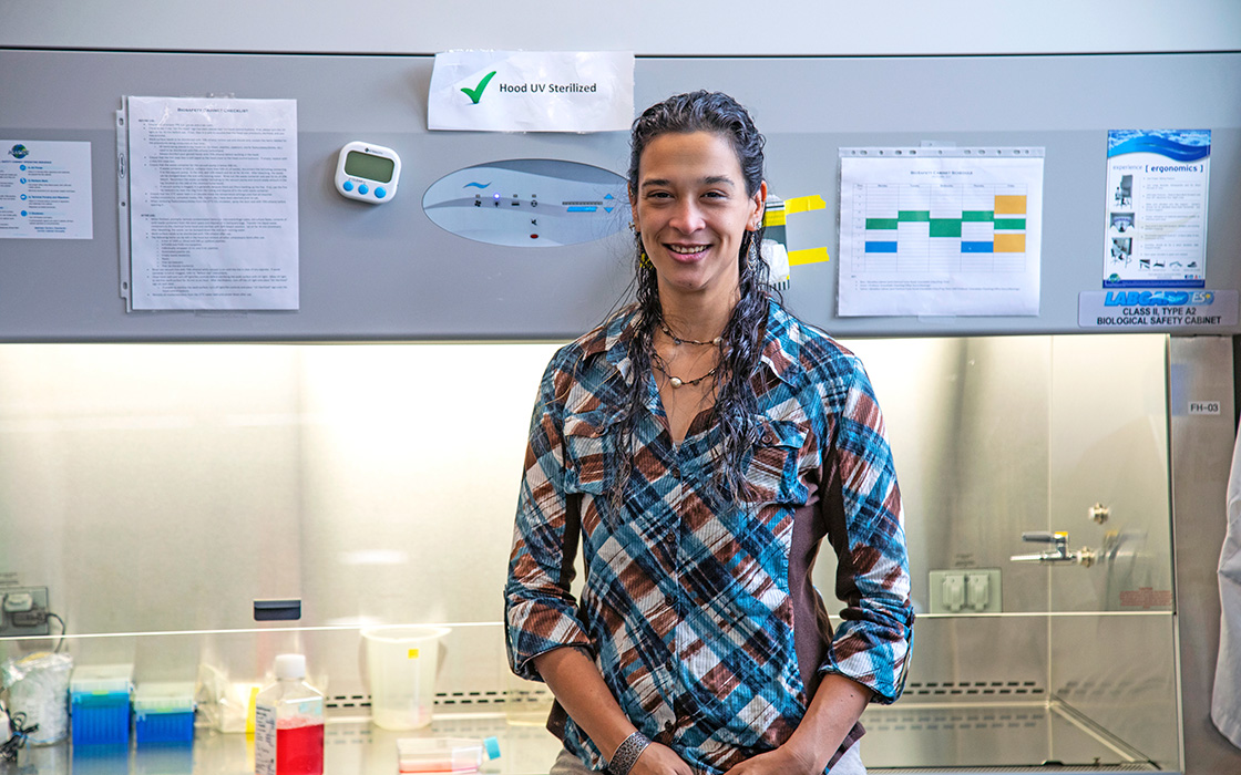 UW Tacoma faculty member Anna Groat Carmona stands in front of a ventilation hood. There are various papers and scientific instruments both behind her and to the side of her. Groat Carmona has long, dark hair and his wearing a plaid shirt. Her hands are crossed at her waist.