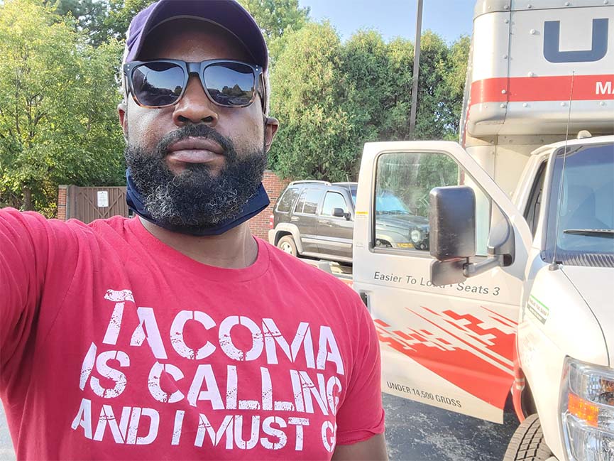 Davon Woodward wearing red t-shirt printed with "Tacoma is calling and I must go," standing in front of U-Haul truck with open door.