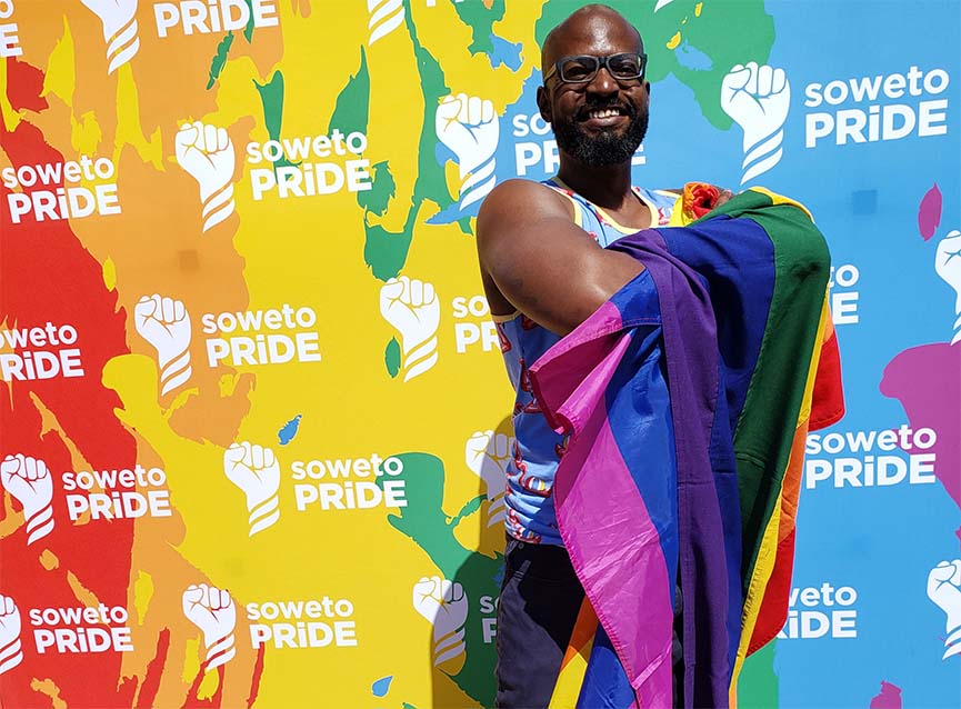 Davon Woodard holding rainbow flag in front of mural with rainbow colors, and upraised fist alongside words "Soweto Pride"