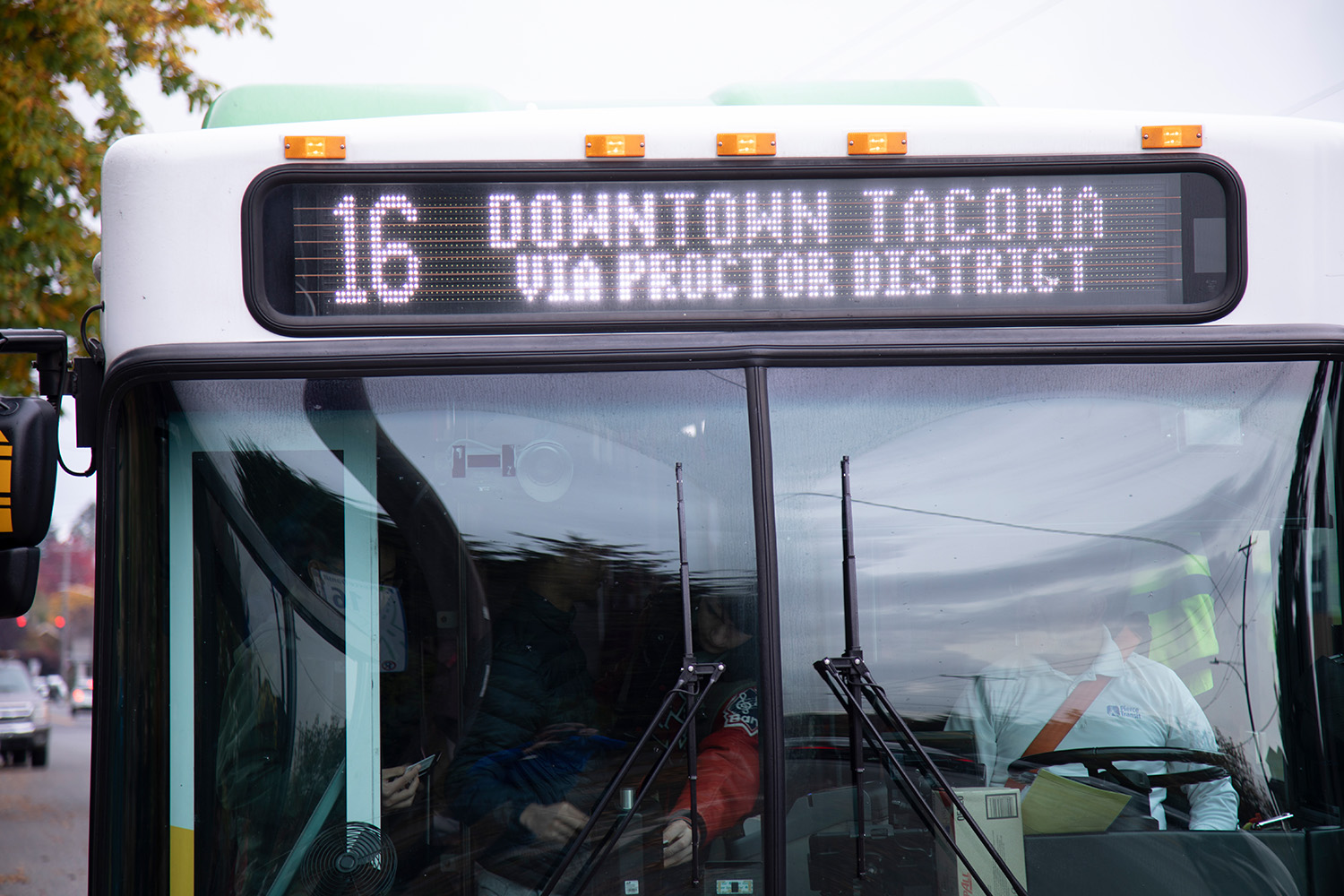 The front of a Pierce Transit bus. There is a glare on the windshield but the driver is partially visible. There is text on the bus reader board that says "16 Downtown Tacoma via Proctor District"