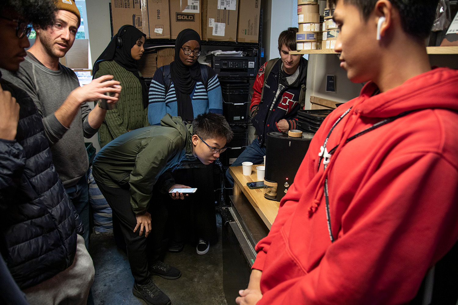 Lander Coffee owner Dustin Johnson talks to a group of students. Johnson is on the left side of the frame. A group of six students form a semi-circle around him. One student has leaned forward to watch as coffee beans get ground in a grinder.