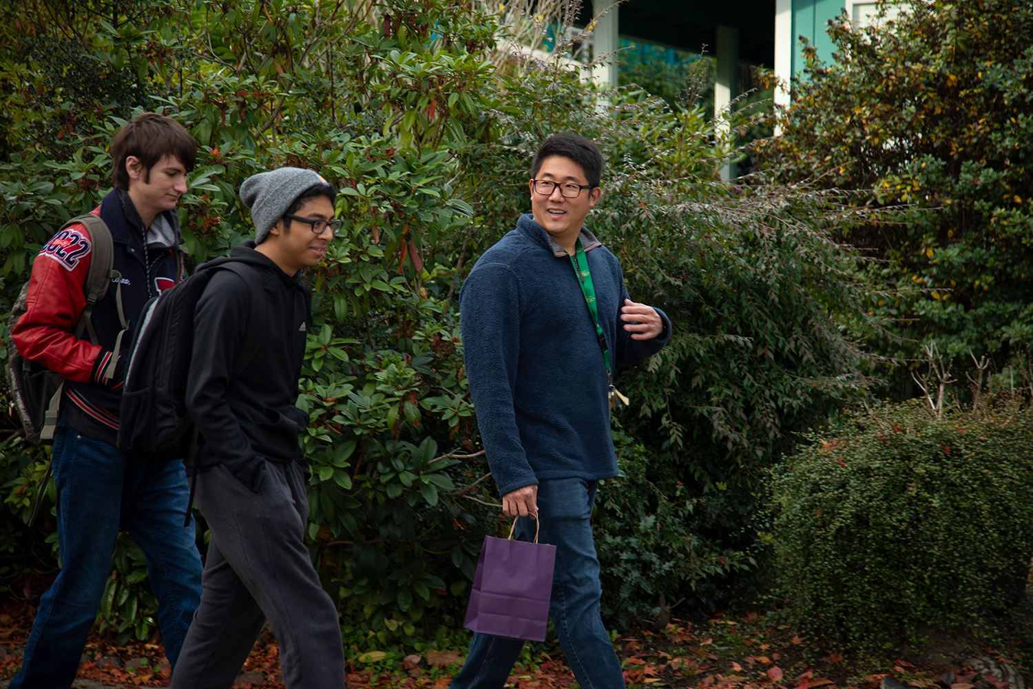 Assistant Professor Seung-Jin Lee talks to two students. The three are walking down a sidewalk. Lee is wearing a dark colored jacket, blue jeans and has short black hair and glasses. The student closest to him is wearing a grey beenie, a dark colored jacket and jeans. The student next to him is wearing a red and gray coat and blue jeans. He has short, brown hair. In the background are different trees and bushes.