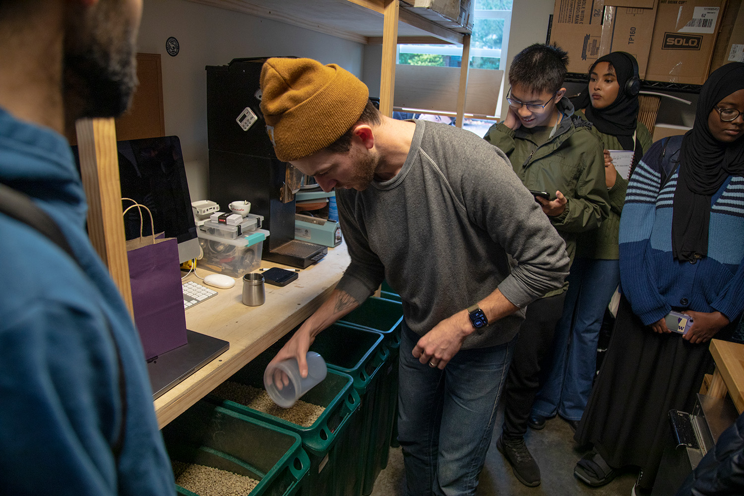 Lander Coffee owner Dustin Johnson uses a plastic cup to scoop out some unroasted green coffee beans from a bin. Johnson is wearing an orange beenie,a gray sweater and blue jeans. There are three students behind him and one to the front and right of where he is standing. There is coffee equipment and boxes off to the side.