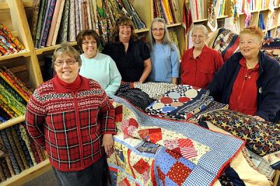 Group portrait of Patti Taylor, '93, and other Operation Mend volunteers posed around various quilts and fabrics.