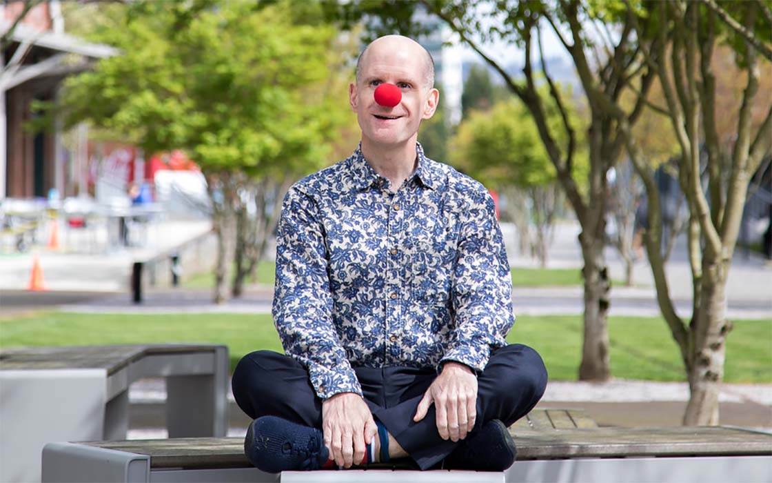 Dr. Luke Perone, UW Tacoma Lecturer, wearing a red clown nose, sitting on a bench on UW Tacoma's Prairie Line Trail.
