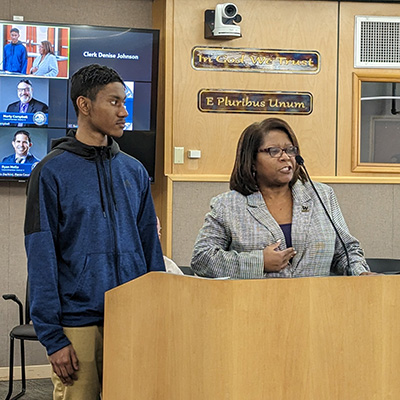 UW Tacoma junior Christian James stands next to Vice Chancellor for Student Affairs Mentha Hynes-Wilson. James is wearing a dark colored coat and khaki pants. Hynes-Wilson is wearing a checkered top. There are TV screens in the background, wood paneling and one window.