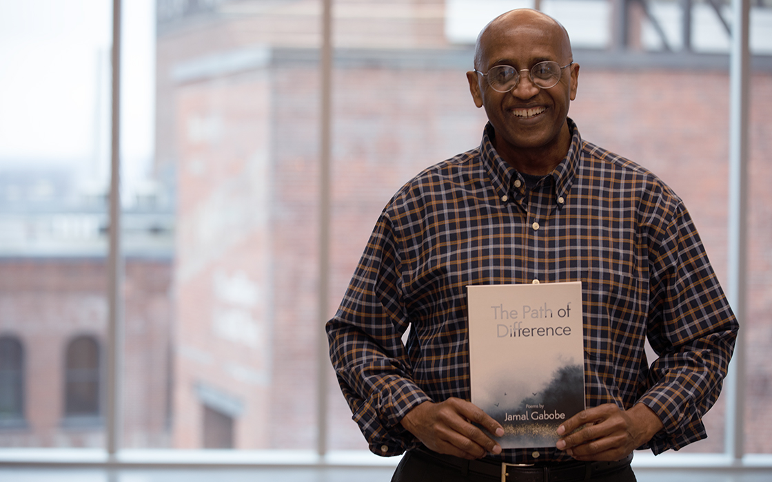 UW Tacoma staff member Jamal Gabobe stands in front of a large window. In the background is a red brick building. Gabobe is holding a copy of his book "The Path of Difference." He is wearing a red plaid shirt and has glasses.