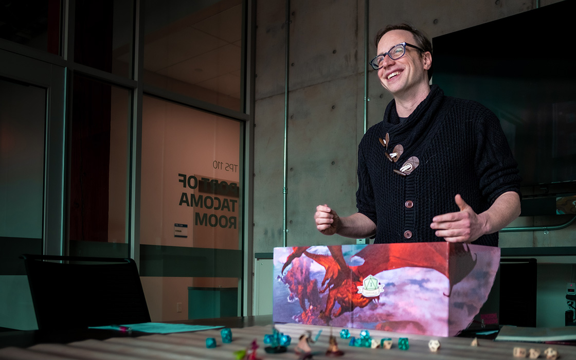 UW Tacoma alumnus Peter Jung stands over a Dungeons & Dragons game board. He has short, brown hair and wears glasses. He is wearing a dark colored sweater and blue jeans. The game board includes miniature figures and a picture of a red dragon in flight.