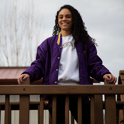 UW Tacoma alumna Amalia Perez stands on a play structure. The structure is brown and made of wood. In the background is a tree with no leaves. Perez is wearing blue jeans, a purple and gold UW coat and a white sweater with the word "Washington" written on it in purple and gold lettering. Perez has long, black, curly hair.